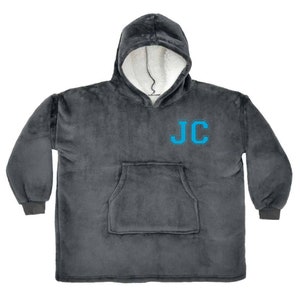 Puede incluir: Una manta gris con capucha y un gran bolsillo en la parte delantera. La manta tiene las iniciales "JC" bordadas en azul en la parte delantera.