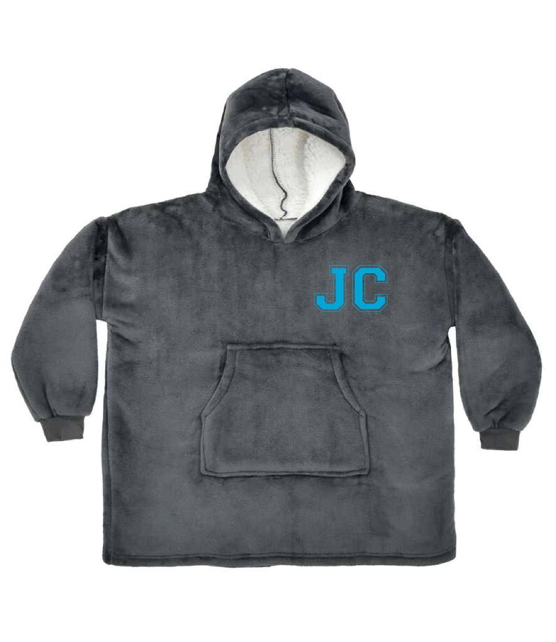 Puede incluir: Una manta gris con capucha y un gran bolsillo en la parte delantera. La manta tiene las iniciales "JC" bordadas en azul en la parte delantera.