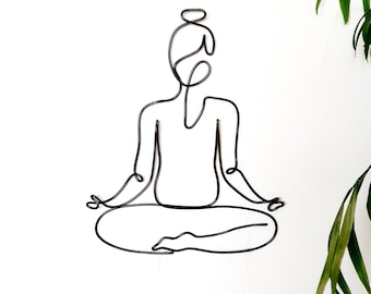May include: A black wire sculpture of a person sitting in a yoga pose. The sculpture is on a white wall.