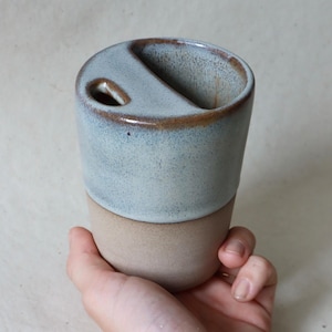 Könnte beinhalten: Ein Keramik-Reisebecher mit zweifarbiger Oberfläche. Der obere Teil ist mit einer glänzenden, gesprenkelten hellblauen Glasur und einem braunen Rand versehen, während der untere Teil ein mattes, unglasiertes beiges Finish aufweist. Der Deckel hat eine Öffnung.
