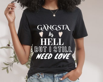 May include: A black t-shirt with white text that reads "Gangsta as Hell But I Still Need Love". The text is surrounded by two pink hearts.
