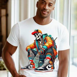 Puede incluir: Camiseta blanca con un gráfico colorido de un esqueleto que lleva una gorra de béisbol, una chaqueta naranja y zapatillas. El esqueleto está sentado con las piernas cruzadas y tiene una cadena de oro alrededor del cuello.