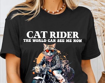 Puede incluir: Camiseta negra con un gráfico blanco de un gato con gafas de sol montando una motocicleta. El texto "Cat Rider The World Can See Me Now" está impreso en la camiseta.