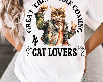 Puede incluir: Camiseta blanca con un gráfico de un gato con gafas de sol y una chaqueta de cuero. El texto "Great things are coming for cat lovers" está impreso en la camiseta.