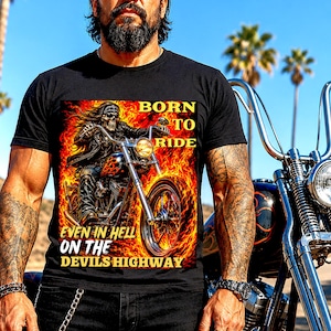 May include: A black t-shirt featuring a graphic print of a skeleton riding a motorcycle engulfed in flames. The text reads BORN TO RIDE EVEN IN HELL ON THE DEVILS HIGHWAY. The design includes vibrant orange and yellow fire patterns surrounding the rider.