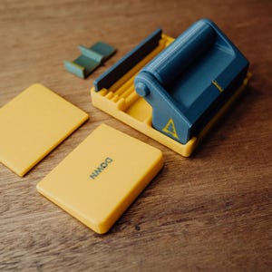 Op de afbeelding: Een blauw en geel 3D-geprint apparaat met een handvat en een geel label met de tekst "DOWN". Het apparaat staat op een houten oppervlak.