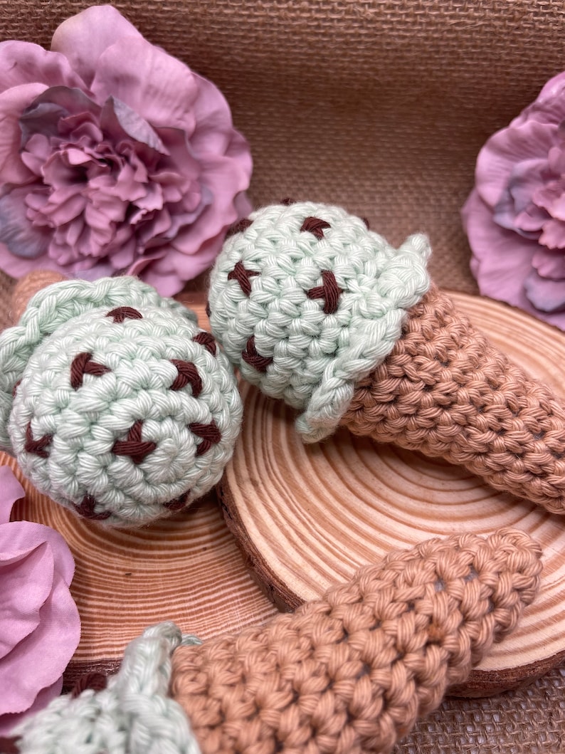Op de afbeelding: Twee gehaakte ijshoorns, een groene met bruine chocolade chips en een bruine, op een houten oppervlak met roze bloemen.