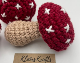 May include: Two crocheted red and white mushroom toys with beige stems. The mushrooms are handmade and have a white cross pattern on the red caps. The text "Klairs Krafts Bespoke Handmade Gifts For You and Your Cat" is on a brown card below the mushrooms.