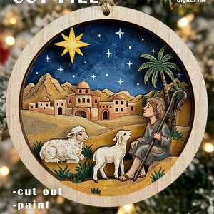 May include: A circular wooden ornament featuring a multi-layered nativity scene with a shepherd, two sheep, a desert town, a palm tree, and a bright yellow star against a dark blue starry sky. Text includes LASER CUT FILE and digital file.