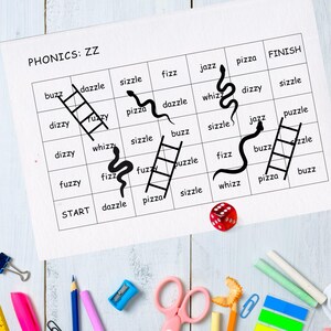 May include: A phonics game board featuring the text "PHONICS: ZZ" and words such as "buzz" and "pizza." Black snakes and ladders are drawn on the board. A red die and stationery items are scattered around the board.
