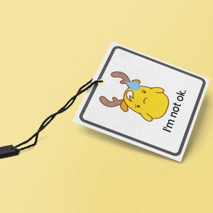 Puede incluir: Una etiqueta cuadrada blanca con un borde negro y una ilustraci&oacute;n de dibujos animados de un personaje amarillo con cuernos y una l&aacute;grima azul. El texto "I'm not ok." est&aacute; impreso en negro en la etiqueta.