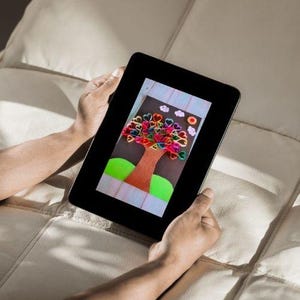 May include: A tablet showing a handmade artwork of a tree. The tree features a brown trunk, green grass, and a crown of heart-shaped leaves in multiple colours. The background is light blue with white clouds and a red flower.