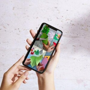 May include: A smartphone screen shows a crafting tutorial with colourful paper cutouts, including a green Christmas tree, butterflies, and geometric shapes. The phone is held in a person's hands against a neutral background.