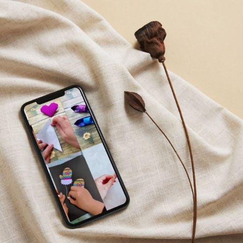 May include: A smartphone screen shows a crafting tutorial with images of handmade items, including a felt heart and decorated mittens. A dried brown flower and seed pod rest on a beige linen surface.