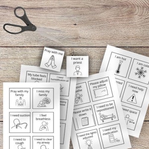May include: A pair of black scissors rests on a light wood surface, alongside several printed sheets of paper. The papers feature black line drawings and text, including phrases like "Pray with me" and "I need my medication."