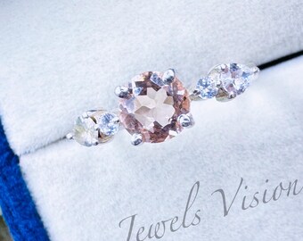 May include: A silver ring with a pink gemstone and two smaller clear gemstones. The ring is in a white box with the text "Jewels Vision" on it.