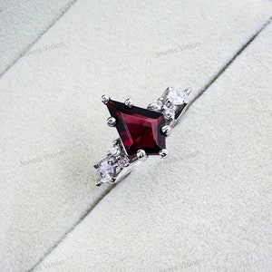 May include: A silver ring featuring a red gemstone in a geometric shape, surrounded by smaller clear gemstones.