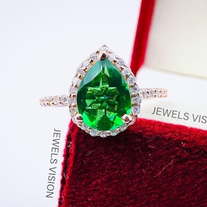 Peut inclure: Une bague en or rose avec une pierre précieuse verte en forme de poire et un halo de petites pierres blanches. La bague est présentée dans un écrin en velours rouge avec le texte "JEWELS VISION" imprimé sur la boîte.