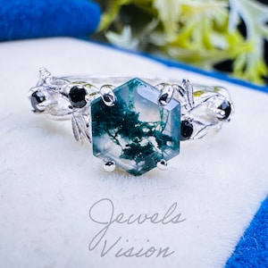 May include: A silver ring with a hexagonal moss agate stone. The ring features black accent stones and leaf-like details on the band. The ring is displayed on a white surface inside a blue box. The text "Jewels Vision" is visible.