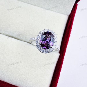 Puede incluir: Un anillo de plata con una gran piedra preciosa ovalada de color púrpura rodeada por un halo de piedras preciosas blancas más pequeñas.