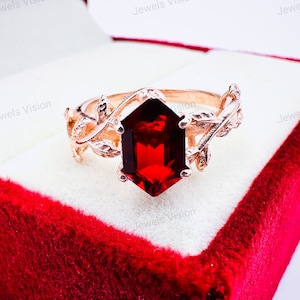 May include: A rose gold ring with a red gemstone set in a hexagonal shape. The ring band is decorated with a delicate leaf design.