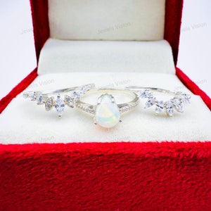 May include: A three-piece ring set featuring a pear-shaped opal center stone and two bands with marquise and round-cut white gemstones. The rings are displayed in a red velvet ring box. The text "Jewels Vision" is visible.