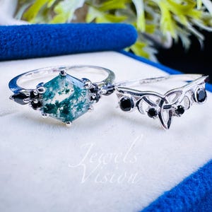Könnte beinhalten: Zwei Silberringe. Ein Ring hat einen sechseckigen Moosachatstein mit schwarzen Akzenten. Der zweite Ring hat ein filigranes, offenes Design mit schwarzen Steinen. Die Ringe werden in einer blauen Samtbox präsentiert.