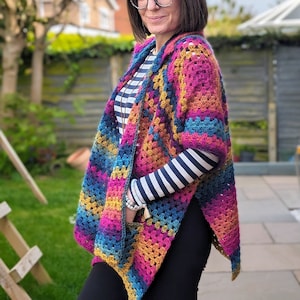 May include: A woman wearing a colorful crocheted poncho with a striped shirt underneath. The poncho is made with a variety of colors, including pink, blue, yellow, and green.