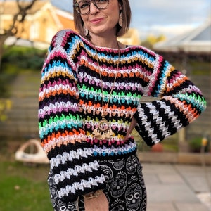 Peut inclure: Un pull en crochet rayé coloré à manches longues. Le pull est réalisé avec une variété de couleurs vives, dont le rose, l'orange, le jaune, le vert, le bleu et le violet. Les manches sont rayées de noir et de blanc.