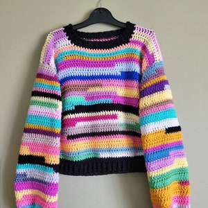 Op de afbeelding: Een kleurrijke gestreepte gehaakte trui met lange mouwen en een ronde hals. De trui is gemaakt met een verscheidenheid aan felle kleuren, waaronder roze, blauw, groen, geel en oranje.