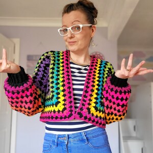 May include: A rainbow crochet top with black trim and long sleeves. The top is cropped and has a V-neck. The model is wearing blue jeans.
