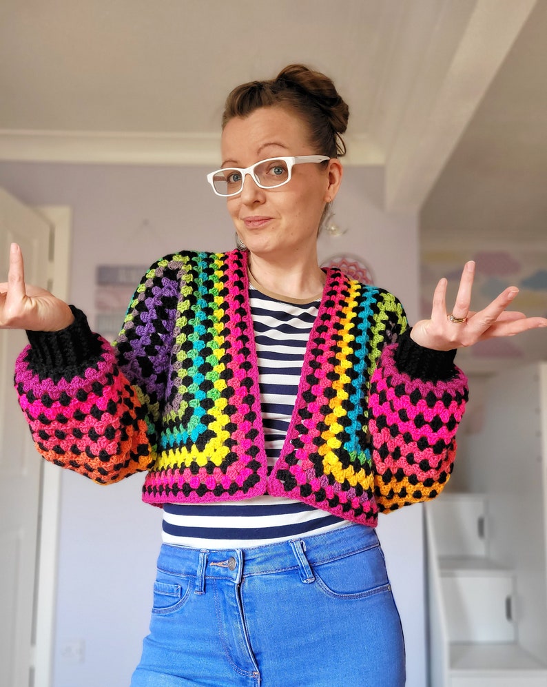 May include: A rainbow crochet top with black trim and long sleeves. The top is cropped and has a V-neck. The model is wearing blue jeans.