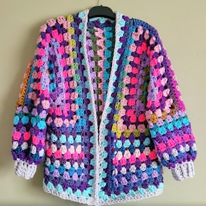 K&ouml;nnte beinhalten: Eine bunte geh&auml;kelte Strickjacke mit einem Granny-Square-Muster in den Farben Lila, Rosa, Blau und Gr&uuml;n. Die Strickjacke hat einen langen, offenen Vorderteil und lange &Auml;rmel.