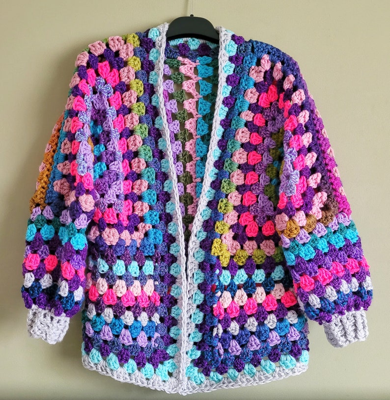 K&ouml;nnte beinhalten: Eine bunte geh&auml;kelte Strickjacke mit einem Granny-Square-Muster in den Farben Lila, Rosa, Blau und Gr&uuml;n. Die Strickjacke hat einen langen, offenen Vorderteil und lange &Auml;rmel.