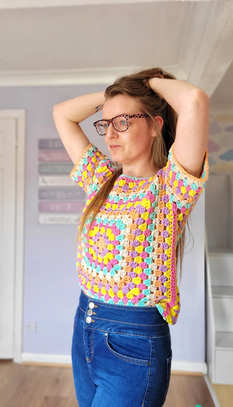 May include: A woman wearing a colorful crochet top with a granny square pattern. The top is made with a variety of bright colors, including pink, yellow, blue, and green. The woman is also wearing blue jeans.