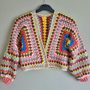 K&ouml;nnte beinhalten: Eine bunte geh&auml;kelte Strickjacke mit einem Granny-Square-Muster in Rot-, Orange-, Gelb-, Gr&uuml;n-, Blau- und Rosat&ouml;nen. Die Strickjacke hat einen V-Ausschnitt und lange &Auml;rmel.