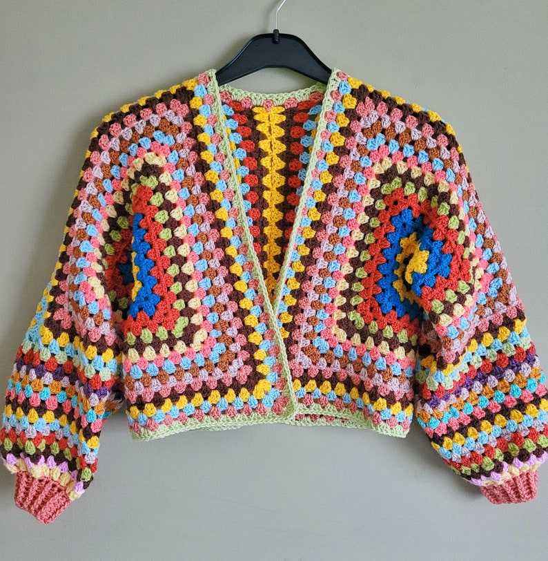 K&ouml;nnte beinhalten: Eine bunte geh&auml;kelte Strickjacke mit einem Granny-Square-Muster in Rot-, Orange-, Gelb-, Gr&uuml;n-, Blau- und Rosat&ouml;nen. Die Strickjacke hat einen V-Ausschnitt und lange &Auml;rmel.