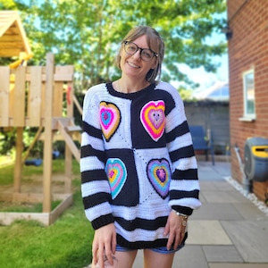 Puede incluir: Una mujer lleva un suéter a rayas blancas y negras con corazones de ganchillo de colores. El suéter tiene un patrón cuadrado con un corazón en cada cuadrado.