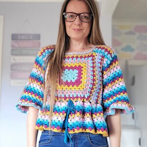 May include: A colorful crocheted top with a square pattern in the center and a tie at the waist. The top is worn with blue jeans.