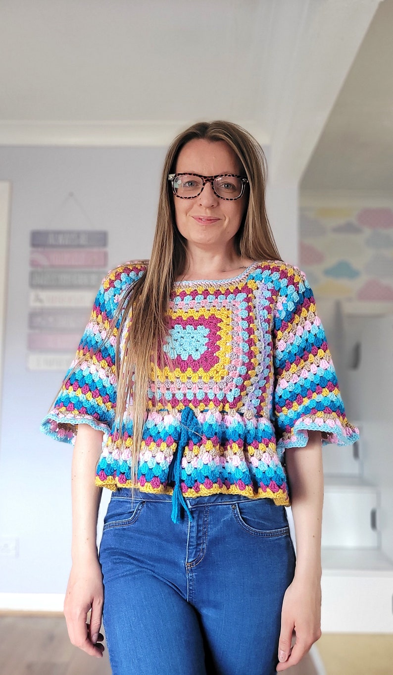 May include: A colorful crocheted top with a square pattern in the center and a tie at the waist. The top is worn with blue jeans.