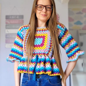 May include: A woman wearing a colorful crocheted top with a blue tassel. The top has a square pattern in the center and is layered with stripes of pink, blue, yellow, and green. She is also wearing blue jeans.