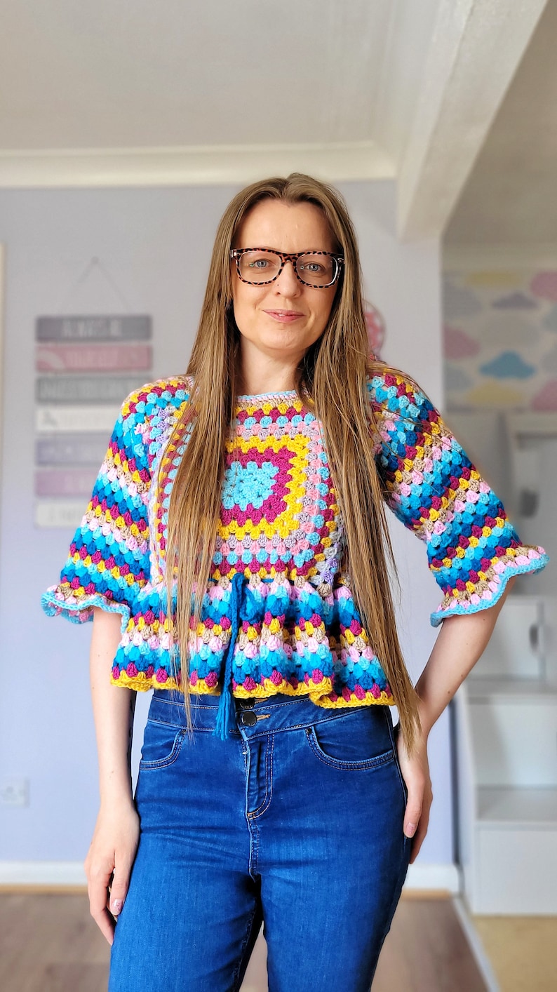 May include: A woman wearing a colorful crocheted top with a blue tassel. The top has a square pattern in the center and is layered with stripes of pink, blue, yellow, and green. She is also wearing blue jeans.