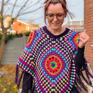 May include: A colorful, handmade crochet poncho with a vibrant, circular pattern in shades of purple, pink, orange, green, and blue. The poncho has a fringed hem and a round neckline. The design features a central mandala-like motif.
