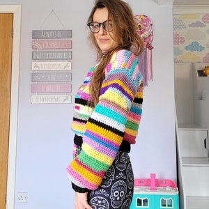 May include: A woman wearing a colorful striped crochet sweater with long sleeves and a black and white skull print leggings. The sweater has a rainbow of colors including pink, yellow, green, blue, and purple. The sign behind her reads "Always be yourself unless you can be a unicorn, then always be a unicorn."