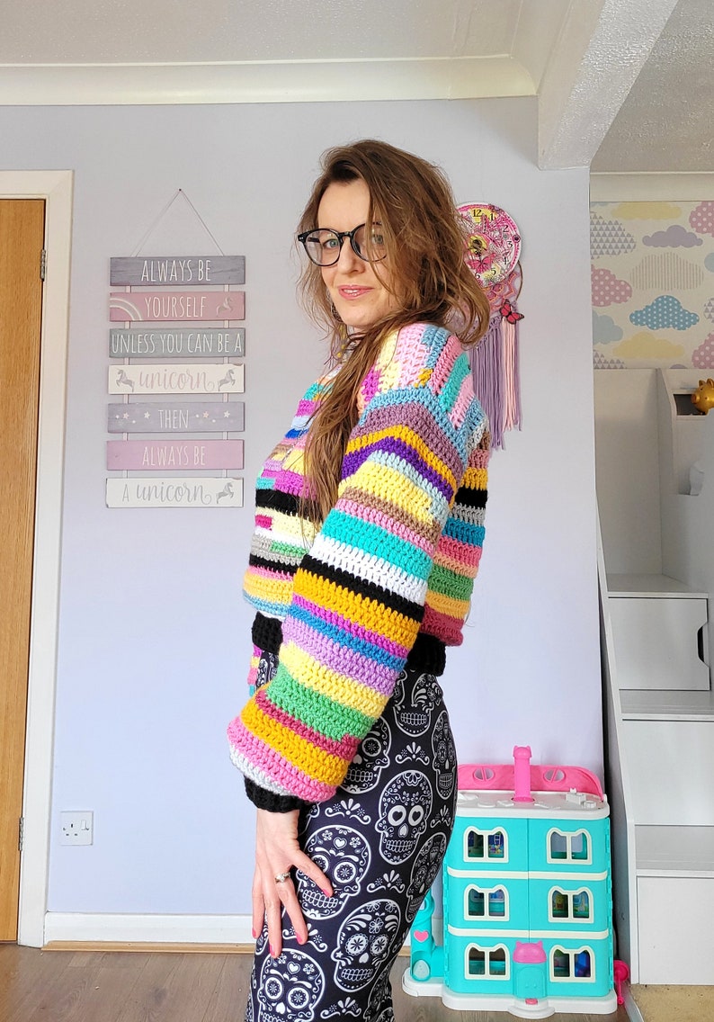 May include: A woman wearing a colorful striped crochet sweater with long sleeves and a black and white skull print leggings. The sweater has a rainbow of colors including pink, yellow, green, blue, and purple. The sign behind her reads "Always be yourself unless you can be a unicorn, then always be a unicorn."