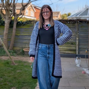May include: A woman wearing a black and white houndstooth cardigan with a black trim, blue jeans, and a colorful necklace.