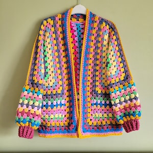 May include: A colorful crocheted cardigan with a granny square pattern. The cardigan has a long, open front and long sleeves. The cardigan is made with a variety of colors, including pink, blue, yellow, green, and purple.
