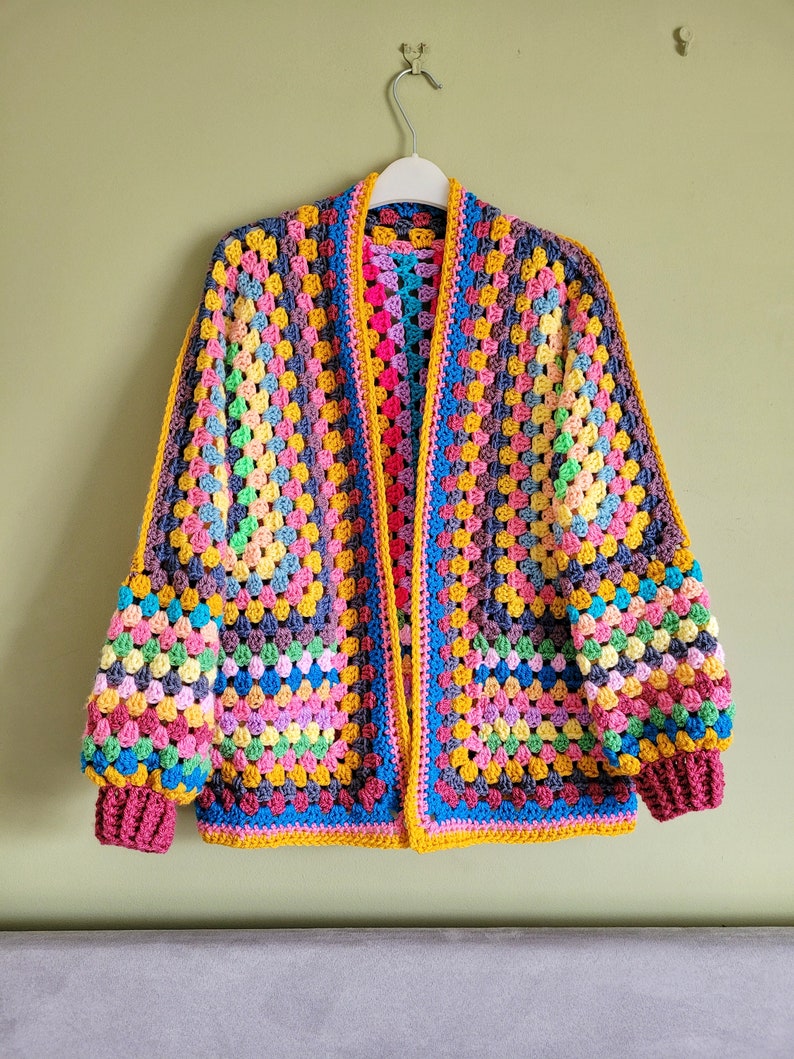 May include: A colorful crocheted cardigan with a granny square pattern. The cardigan has a long, open front and long sleeves. The cardigan is made with a variety of colors, including pink, blue, yellow, green, and purple.