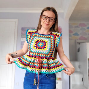 May include: A colorful crochet top with a square design and a ruffled bottom. The top has a tie at the waist and is made with a variety of bright colors, including red, yellow, blue, green, and purple.