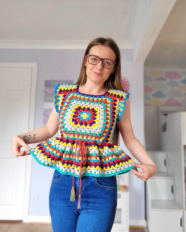 May include: A colorful crochet top with a square design and a ruffled bottom. The top has a tie at the waist and is made with a variety of bright colors, including red, yellow, blue, green, and purple.